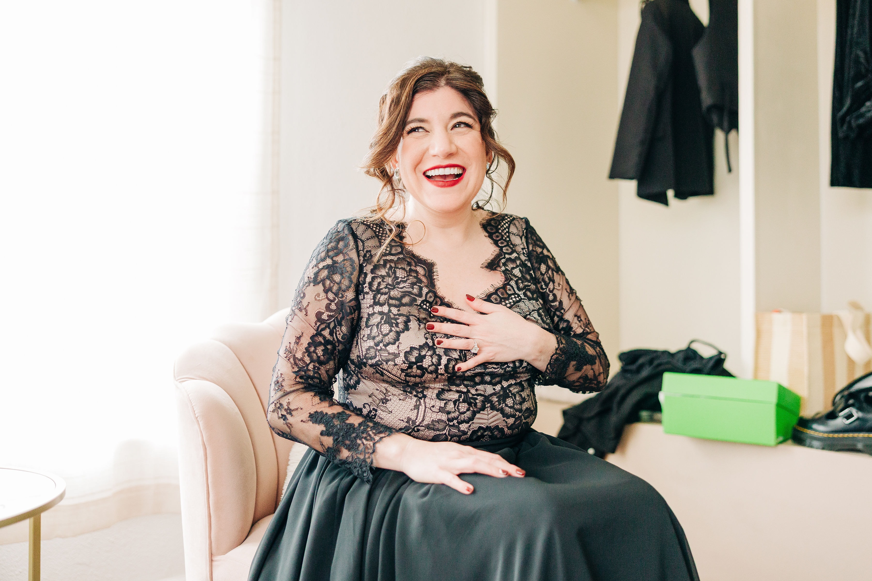 Jenn sitting in the bridal suite with a huge smile on her face, laughing at a joke her friend just made. The bright natural light contrasts with her black lace dress.