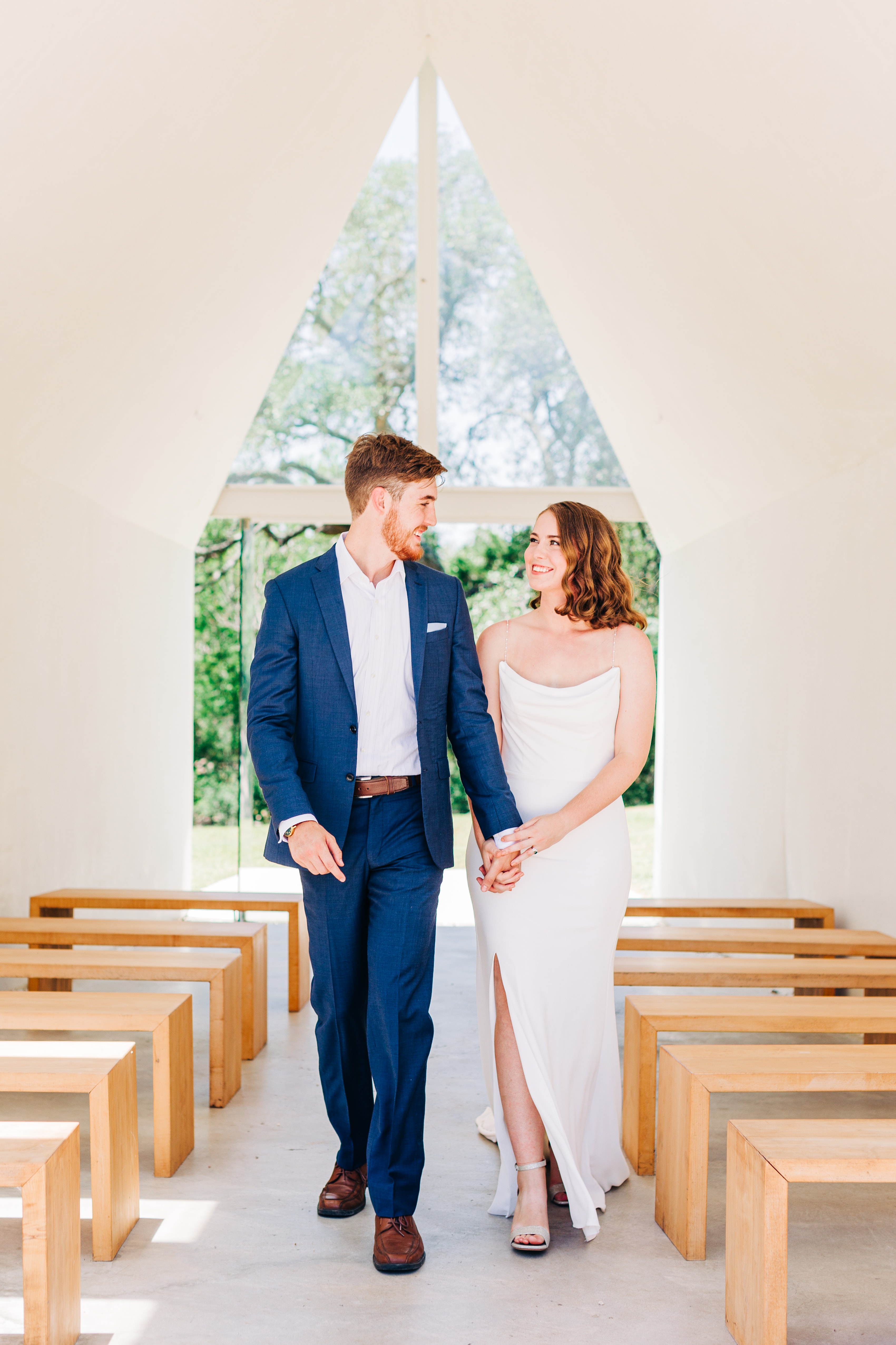 A couple walks hand in hand down the aisle of lincoln chappel, locking eyes and smiling with the clean minimalist walls and benches of the chapel.