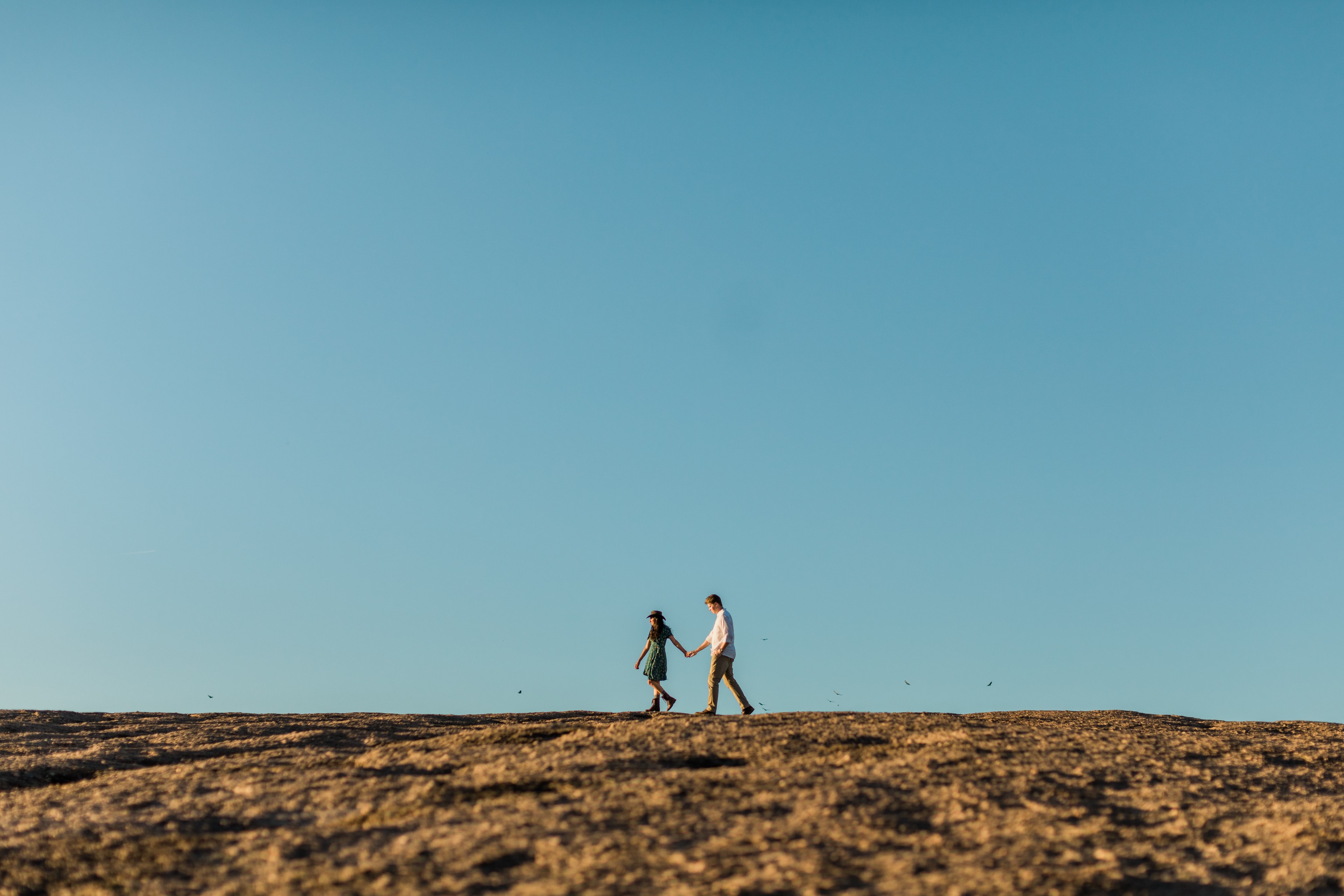 Couple walking across the stone surface of Enchanted Rock. The wide landscape and blue skies surround them as birds fly in the distance.