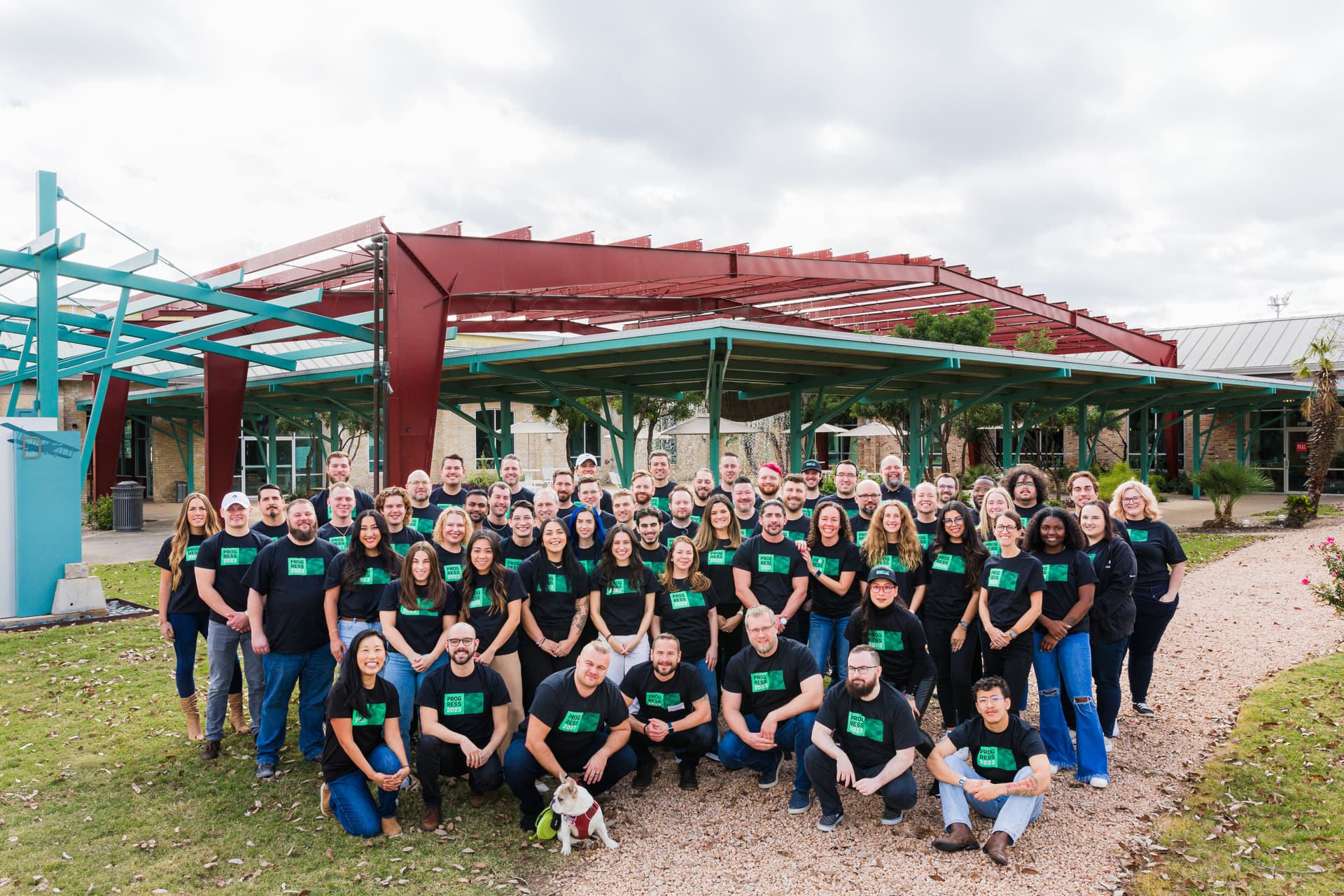 A large group portrait of a company wearing coordinated team shirts in natural light, in front of their office. the employees gather around in a relaxed and confident matter, smiling and connecting with eachother.