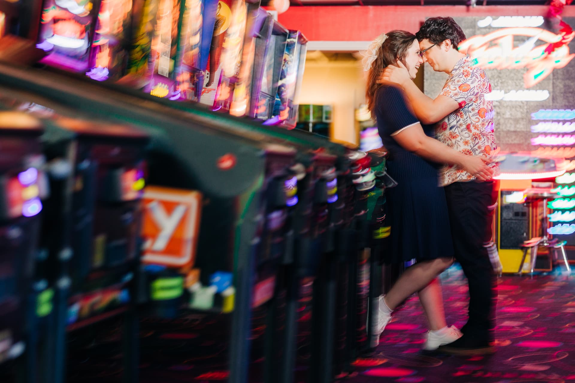 Couple embracing by a vintage arcade game together at Pinballz, bathed in colorful neon light with genuine laughter on their faces
