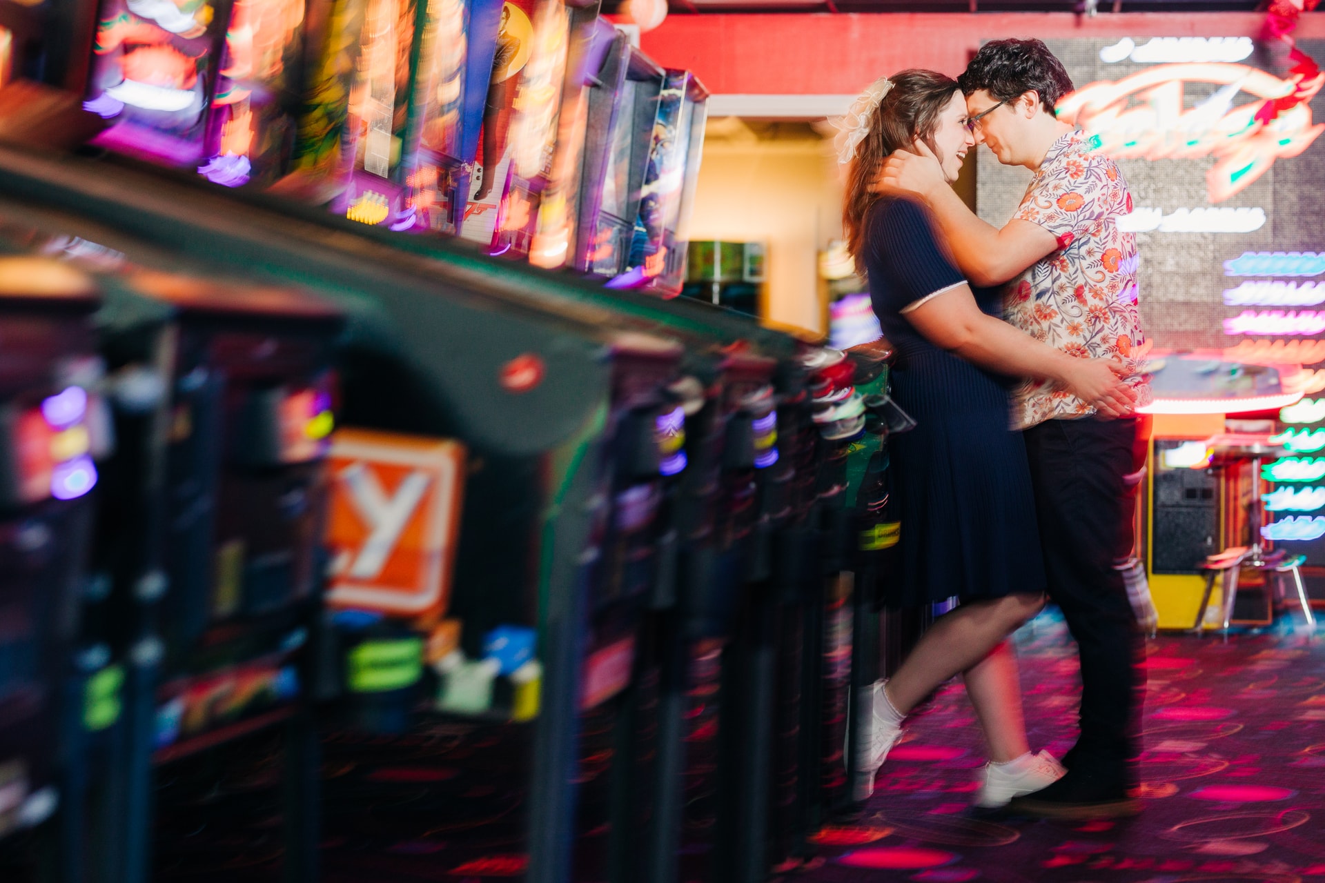 Couple embracing by a vintage arcade game together at Pinballz, bathed in colorful neon light with genuine laughter on their faces
