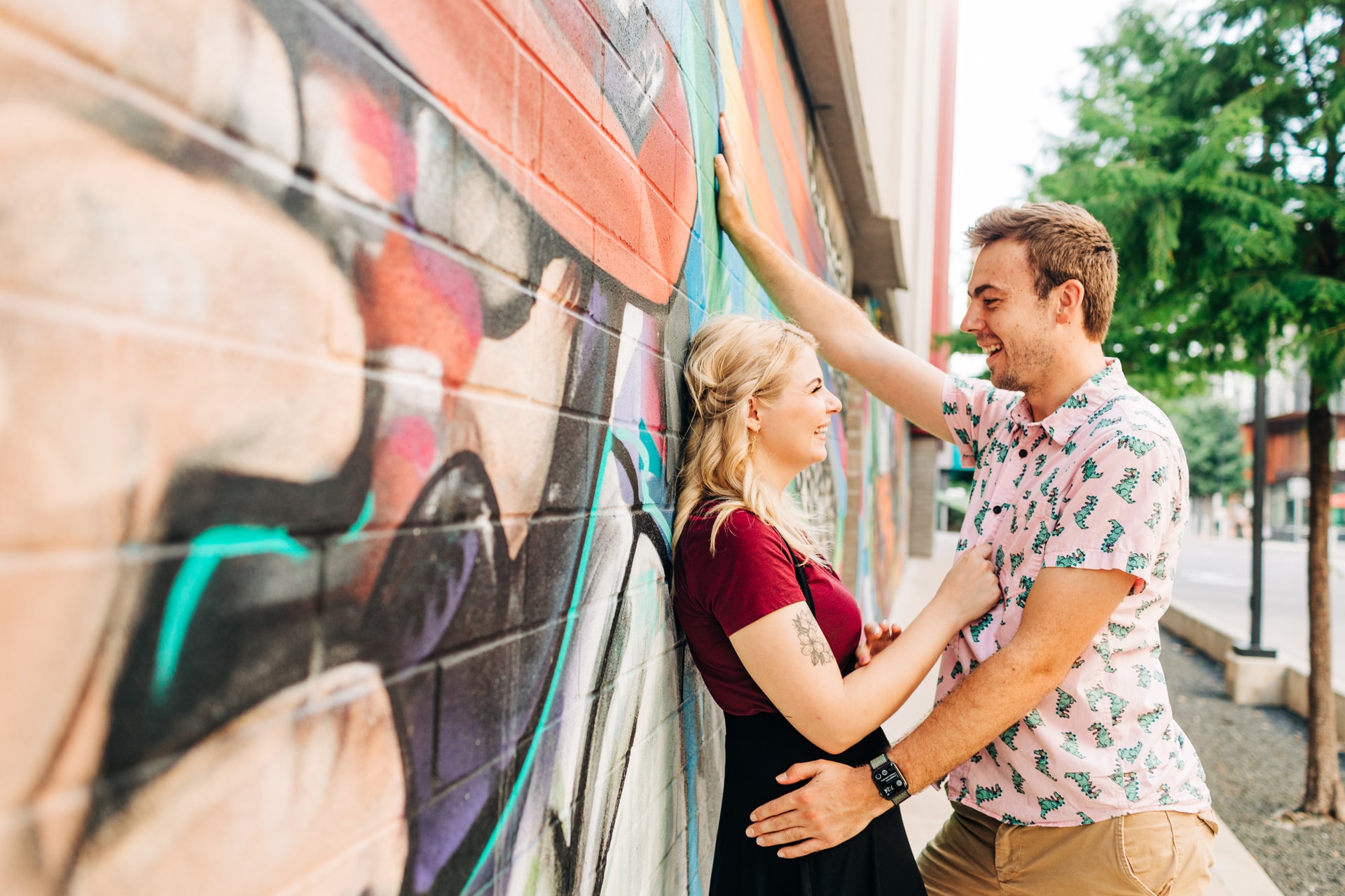Couple cuddled up to eachother against a graffiti wall near the South Congress Alamo Drafthouse area, the lighting is natural and their smiles are authentic