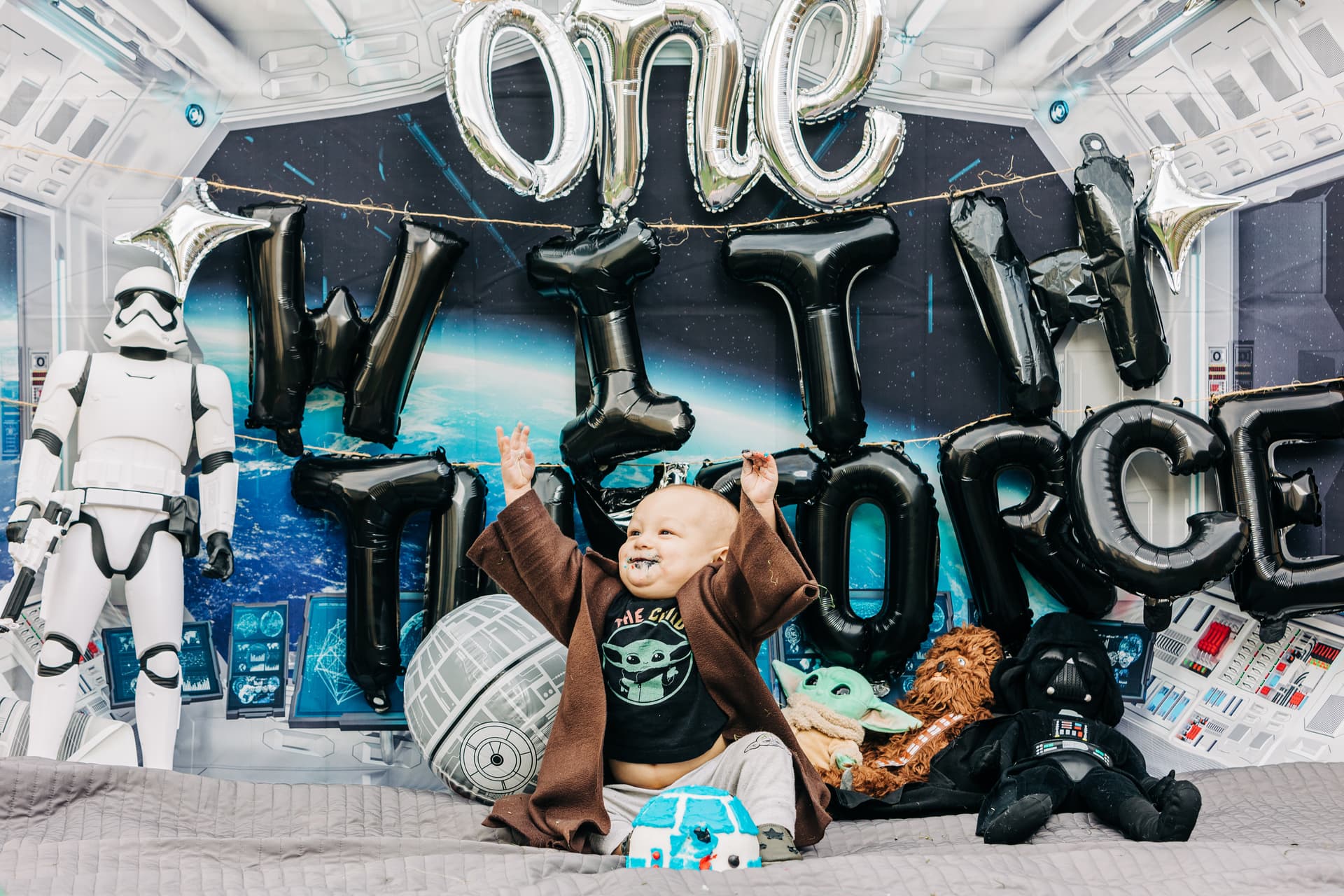 One-year-old elbow-deep in birthday cake frosting during a cake smash session — wide-eyed, messy-faced delight with colorful cake crumbles everywhere and family watching in the background