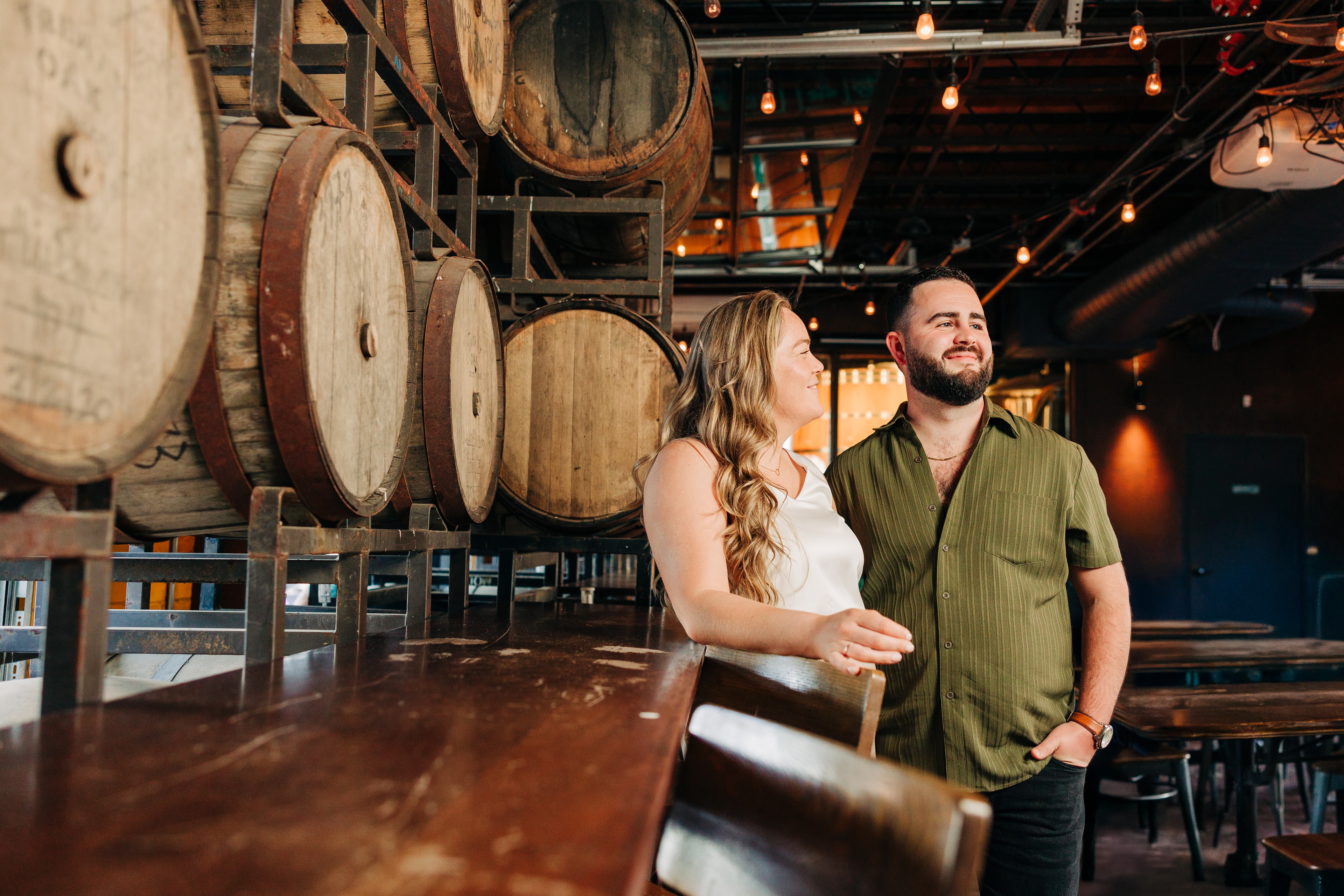 Engaged couple wearing complementary earth tones — one in muted green and the other in cream — coordinated but not matching, standing close together in warm golden light