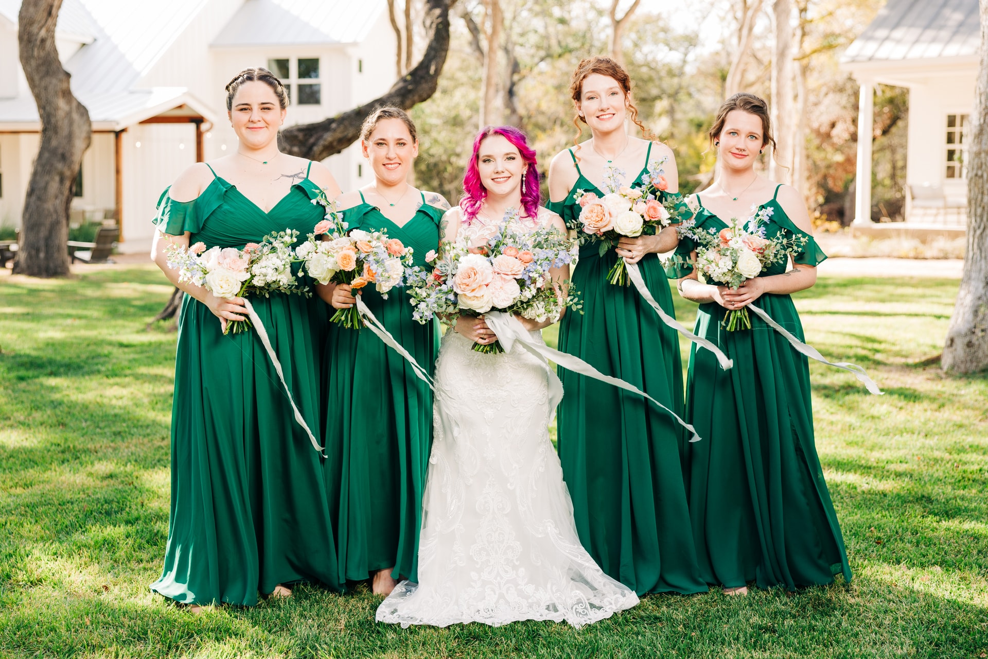 a bridal party joyfully smiles around the bride with pink and purple hair as she anticipates her walk down the aisle, the group is covered in the warm glow of natural light with white cottages behind them.