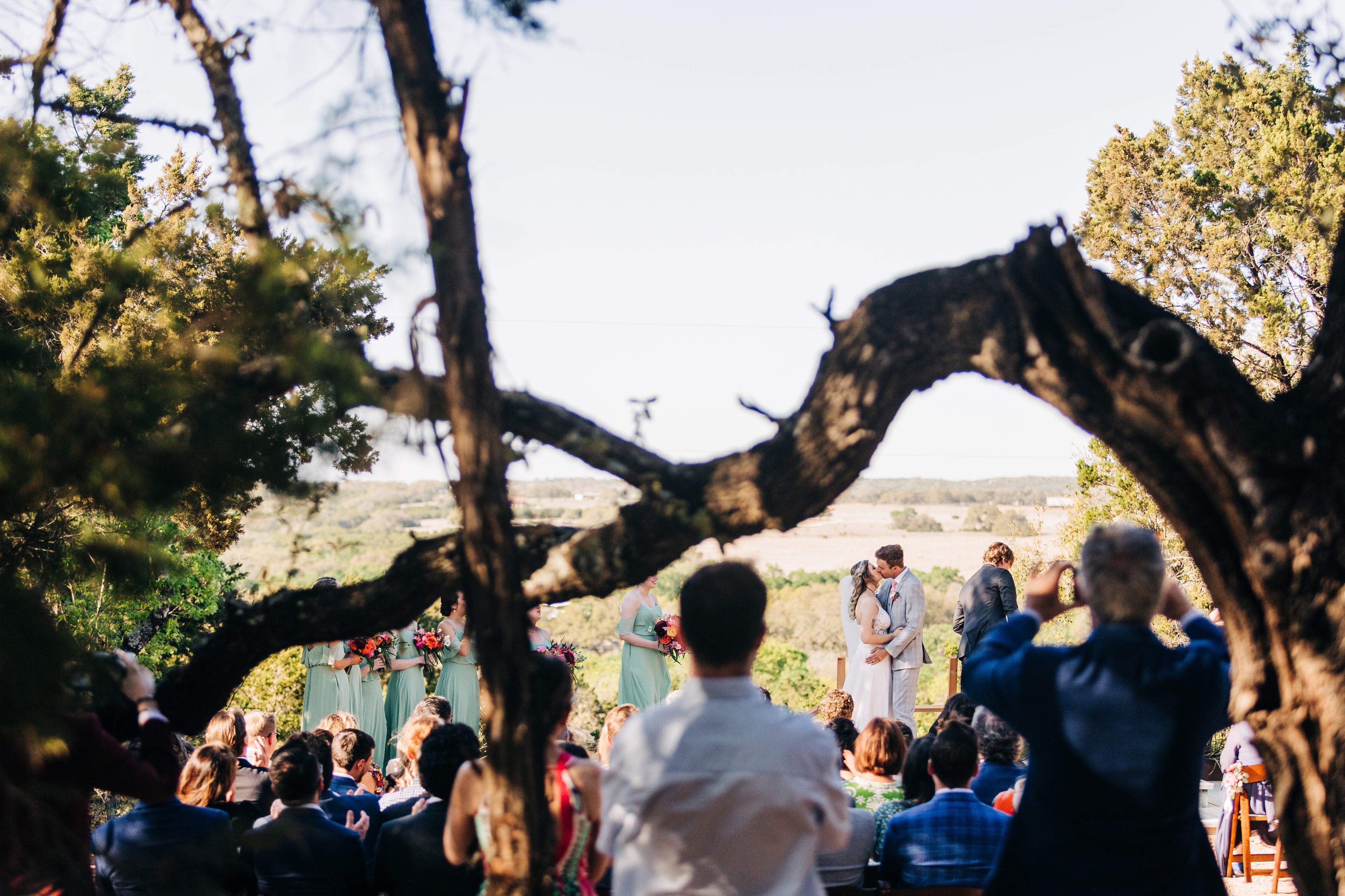 Wedding ceremony at Lucky Arrow Retreat with wide Hill Country vista behind the couple, guests seated along the winding walkway, framed by a large oak tree