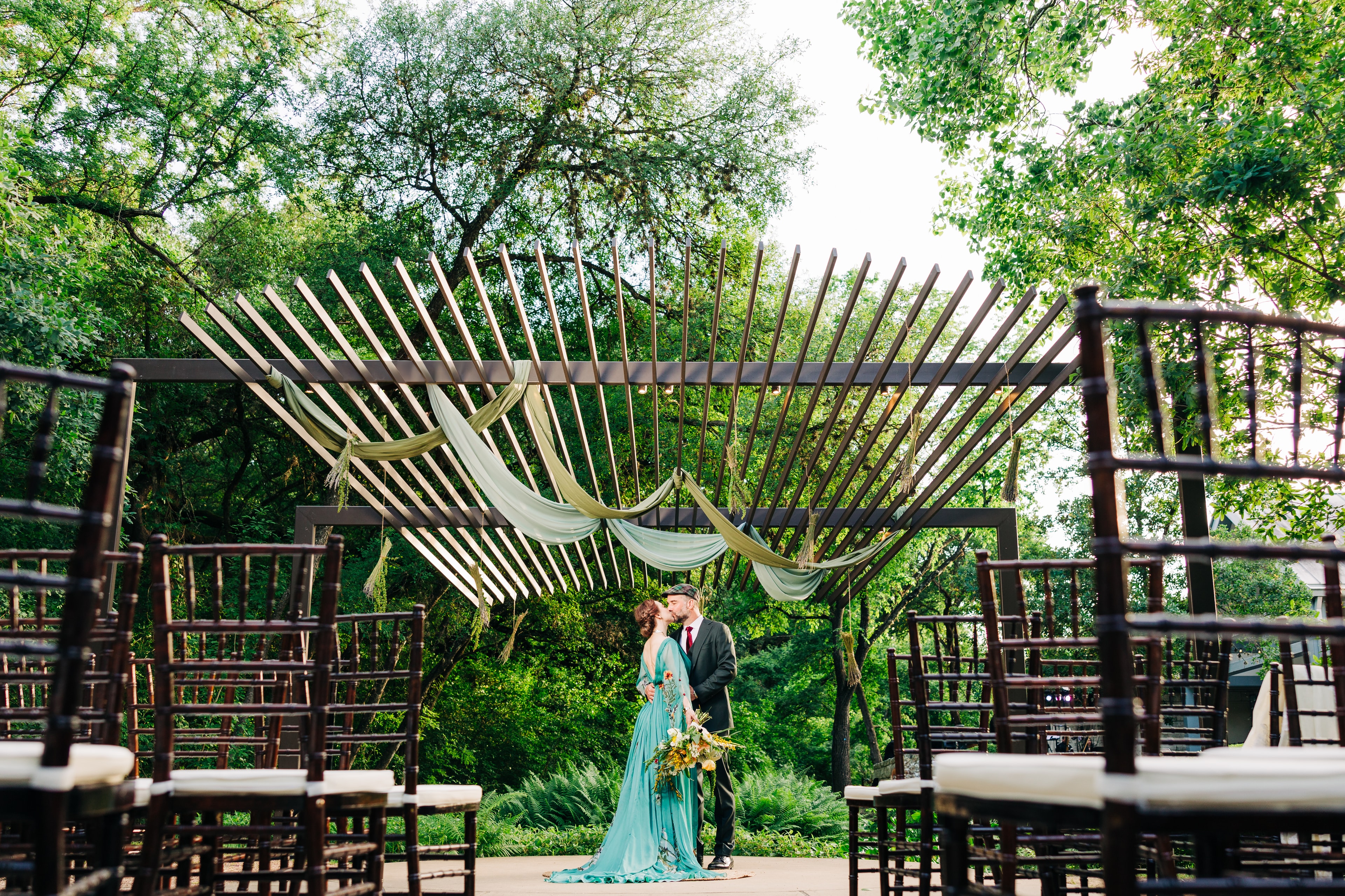 A wide angle portrait of a couple at the Umlauf sculpture garden ceremony arch, framed by the ceremony chairs and embracing. the couple is wearing alternative wedding attire, with the bride in green and the groom wearing a hat.